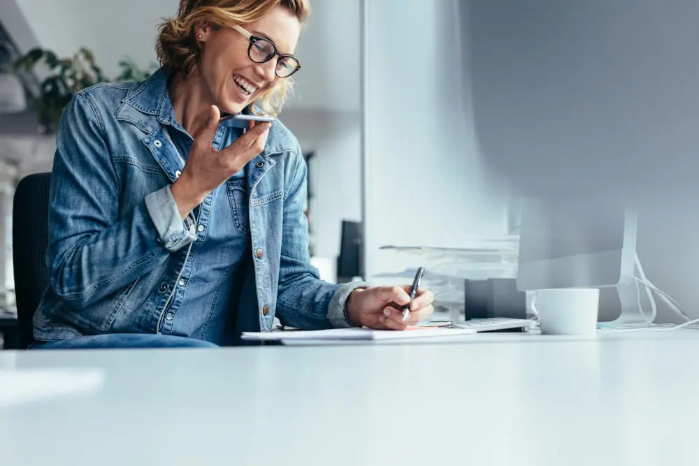 Woman working at her desk at home. She's writing and talking on the phone.